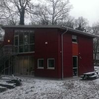 Ein rotes Haus im Schnee, im Hintergrund steht ein Baum, der die winterliche Landschaft ergänzt.