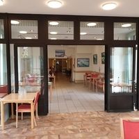 Ein Restaurant mit Tischen und Stühlen vor großen Fenstern, die viel Tageslicht hereinlassen.